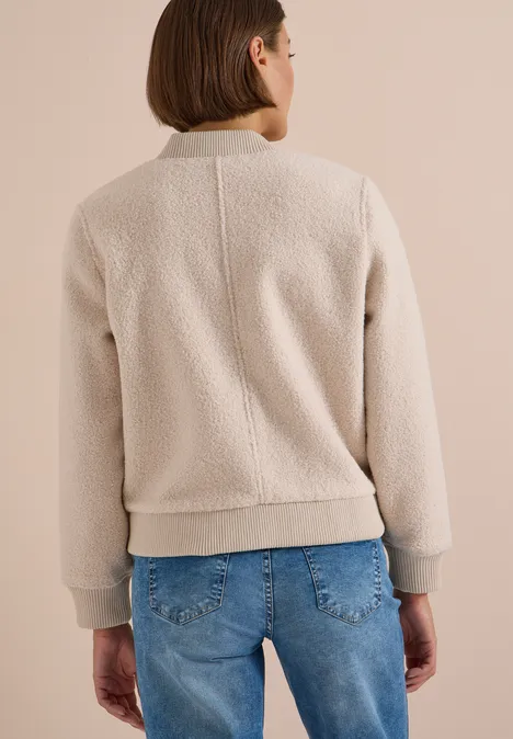 Achteraanzicht van persoon in beige fleece bomberjack met groene boorden en blauwe spijkerbroek voor beige achtergrond.
