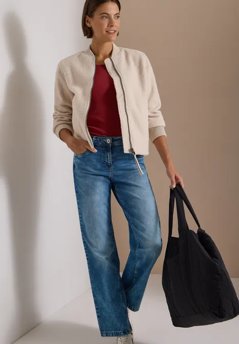 Vrouw draagt beige bomberjack, rood topje, jeans en zwarte tas, nonchalant leunend tegen witte muur.