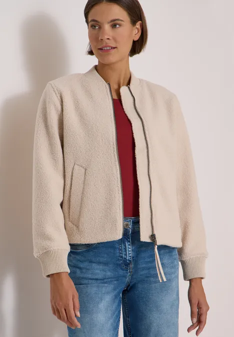 Vrouw in beige teddyjack, rood shirt en blauwe spijkerbroek, staand voor een neutrale achtergrond.