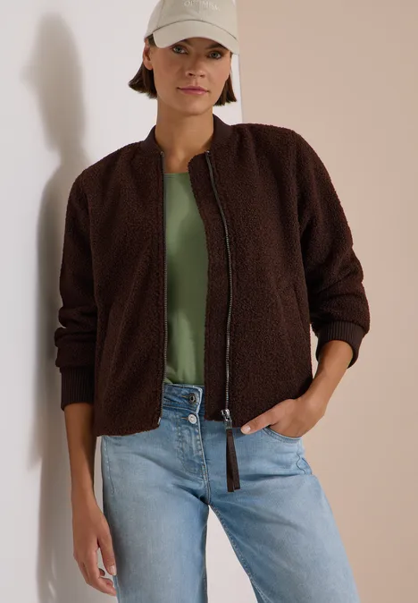 Frau lehnt an Wand, trägt braune Teddy-Jacke, grünes Top, helle Jeans und hellen Cap, lässiger Look