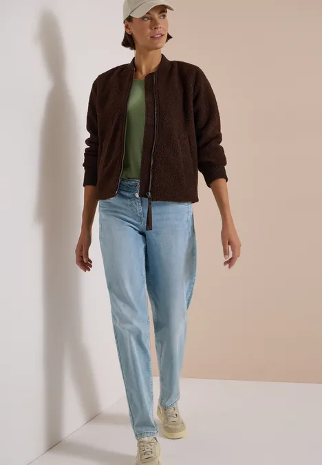 Frau in dunkelbrauner Jacke, grünem Shirt, hellblauen Jeans, Sneakers und beigem Baseballcap vor schlichtem Hintergrund.