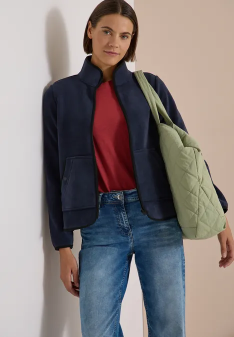 Fleecejacke mit Zipper und Stehkragen dark navy blue Frau posiert vor neutralem Hintergrund mit dunkelblauer Jacke, rotem Shirt, Jeans und hellgrünem Steppbeutel.