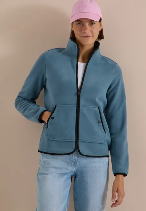Fleecejacke mit Zipper und Stehkragen mountain air blue Frau trägt petrolfarbene Fleecejacke mit Reißverschluss, Taschen, über weißem Shirt, rosa Cap, Jeans.