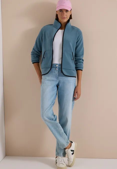 Fleecejacke mit Zipper und Stehkragen mountain air blue Eine Frau steht in Jeans, weißem Shirt, blauem Fleece, rosa Cap und Sneakern vor beige.