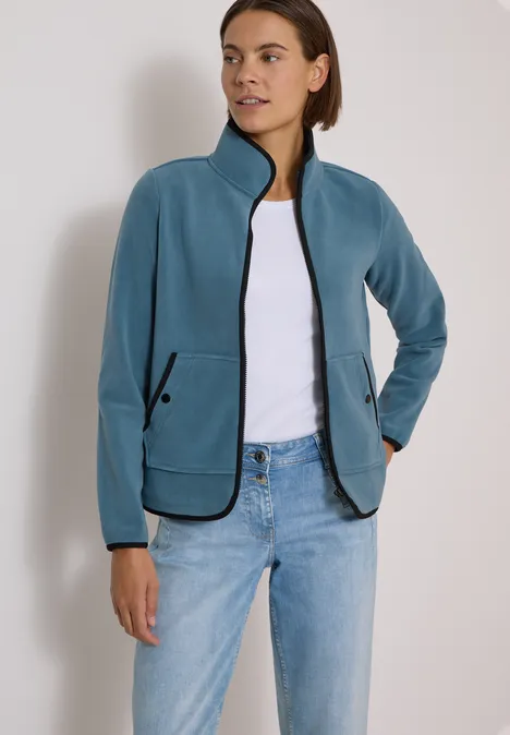 Fleecejacke mit Zipper und Stehkragen mountain air blue Frau trägt türkisblaue Fleecejacke mit Reißverschluss und schwarzen Details, dazu weißes T-Shirt und Jeans.