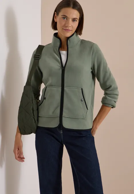 Fleecejacke mit Zipper und Stehkragen shadow khaki Frau trägt grüne Fleecejacke, weißes Shirt, blaue Jeans und grüne Umhängetasche vor hellem Hintergrund.