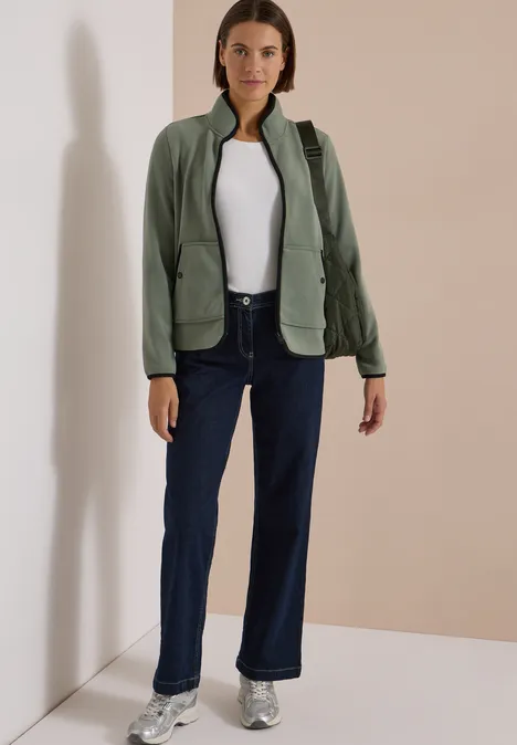 Fleecejacke mit Zipper und Stehkragen shadow khaki Frau steht in Raumecke, trägt dunkle Jeans, weiße Bluse, grüne Jacke und Sneaker.