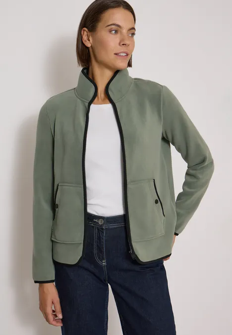 Fleecejacke mit Zipper und Stehkragen shadow khaki Frau trägt olivgrüne Fleecejacke mit Reißverschluss und Taschen, kombiniert mit weißem Shirt und Jeans.
