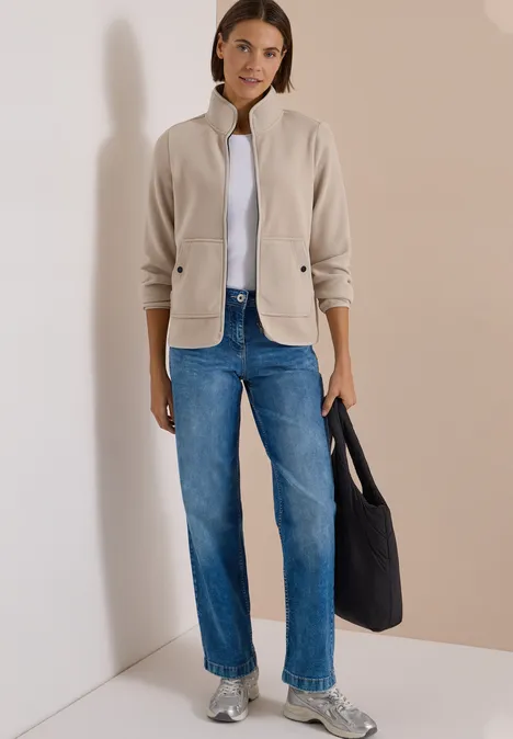 Fleecejacke mit Zipper und Stehkragen powder beige Frau in heller Jacke, weißem Shirt, blauen Jeans und silbernen Sneakern mit schwarzer Tasche vor neutraler Wand.