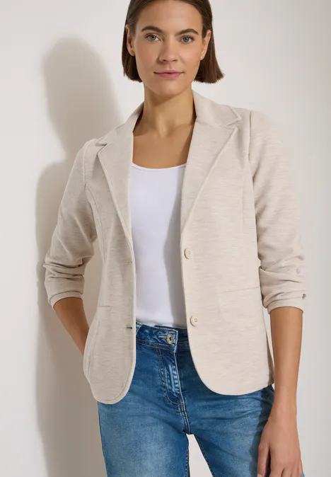 Vrouw in lichtbeige blazer, wit topje en blauwe jeans, ontspannen houding met handen in zakken.