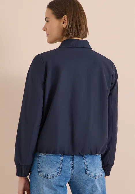 Blouson mit Hemdblusenkragen urban dark blue Rückansicht einer Person in dunkelblauer Jacke und blauer Jeans vor beigem Hintergrund.