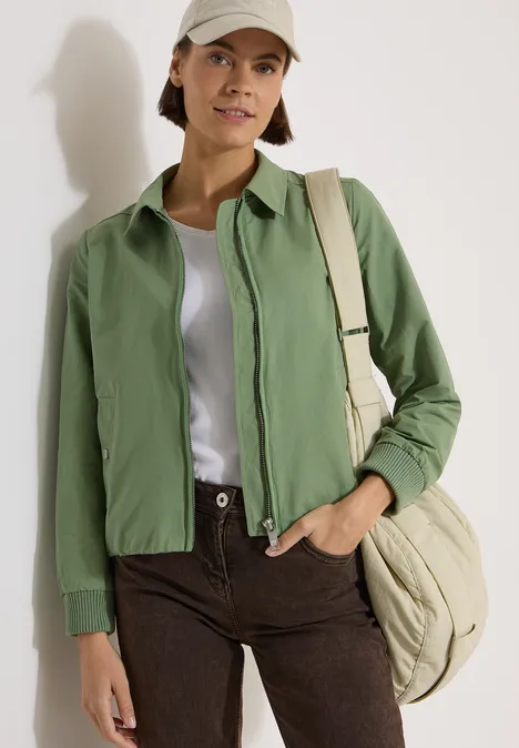 Iemand draagt een groene jas, wit T-shirt, bruine broek en een beige tas.