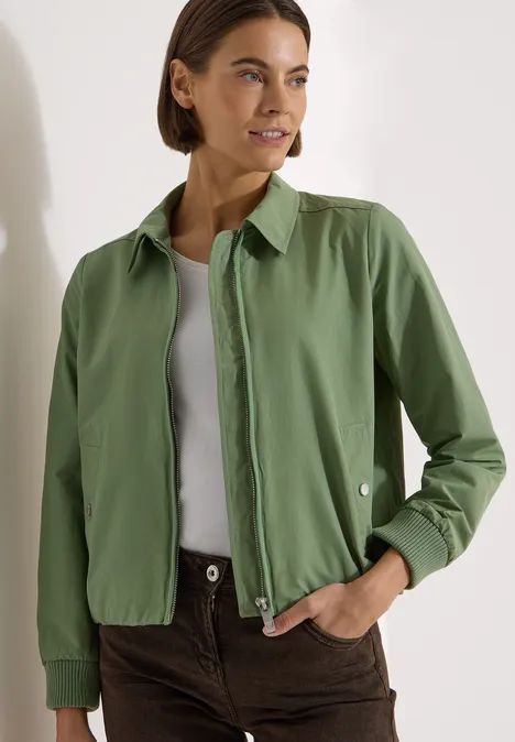 Persoon draagt groene overshirt, wit hemdje en bruine broek in een minimalistische, modieuze look.
