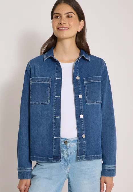 Frau in Jeansjacke und Jeanshose mit weißem Shirt lächelt vor neutralem Hintergrund.