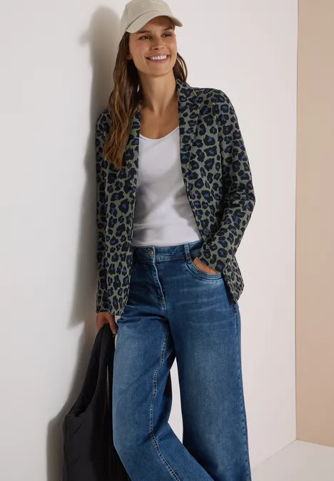 Vrouw leunt ontspannen tegen muur, draagt luipaardblazer, wit shirt, wijde jeans en pet, glimlacht.