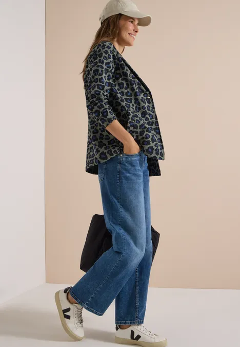 Vrouw met pet draagt motiefblazer, wijde spijkerbroek en witte sneakers voor een beige muur.