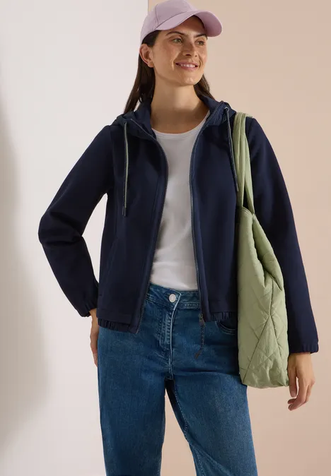 Frau in dunkelblauer Kapuzenjacke, weißem T-Shirt, Jeans und grüner Stepp-Tasche, lächelnd mit lila Cap