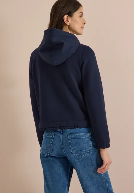 Frau steht vor beigem Hintergrund, trägt dunkelblaue Kapuzenjacke und Jeans, Rückansicht.