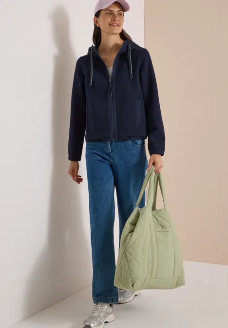 Eine Frau trägt eine blaue Kapuzenjacke, Jeans, Cap und hält eine große hellgrüne, gesteppte Tasche.