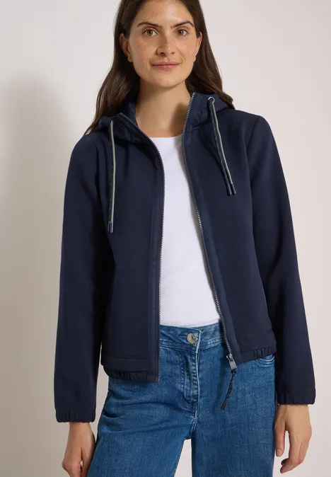 Frau in dunkelblauer Kapuzenjacke über weißem Shirt und blauen Jeans vor hellem Hintergrund.
