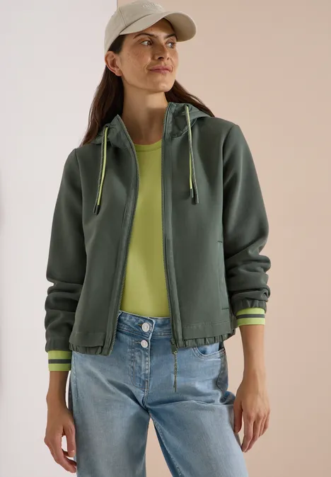Eine Frau in grüner Kapuzenjacke, neonfarbigem Shirt und Jeans vor hellem Hintergrund.