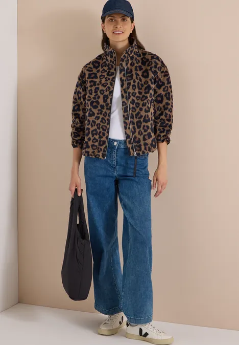 Frau trägt Leopardenjacke, weiße Bluse, blaue Jeans, Sneaker, Baseballcap und hält eine dunkelblaue Tasche.