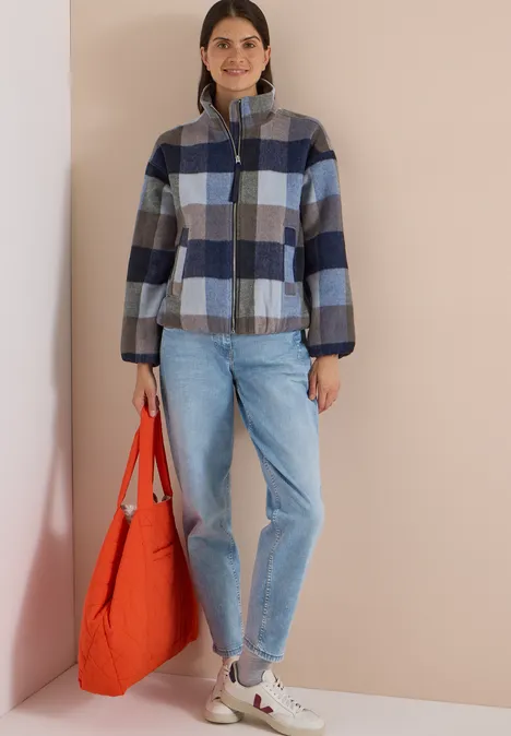 Frau in blau-grauer karierten Jacke, Jeans und weißen Sneakern hält eine orangefarbene Tasche.