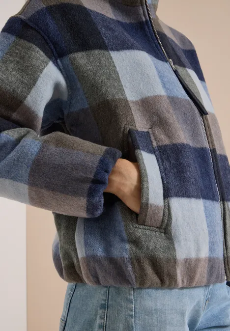 Person trägt blau-karierte Jacke mit Reißverschluss und Tasche, dazu Jeans.