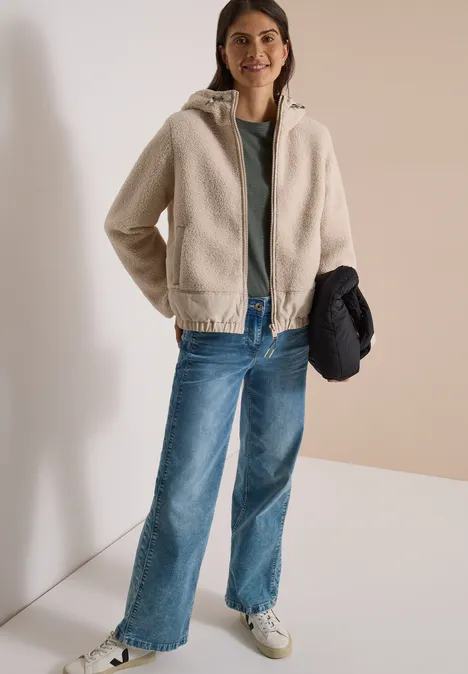 Frau in beiger Teddyjacke, grünem Shirt, blauen Jeans und weißen Sneakern, hält dunkle Jacke in der Hand.