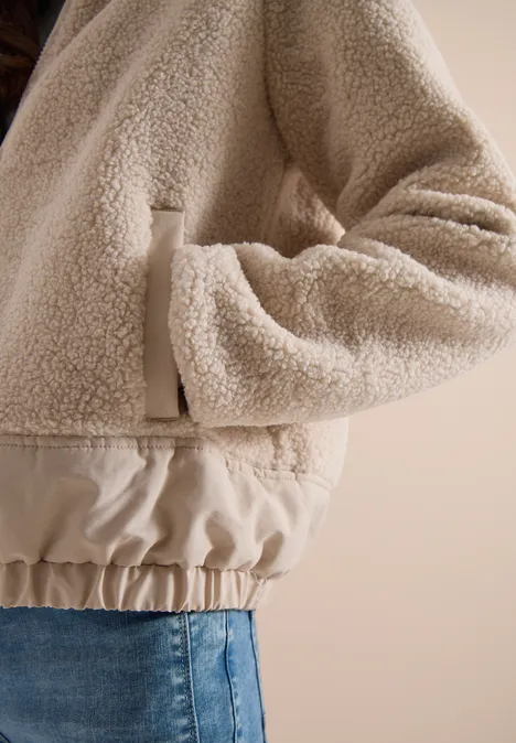 Beige Teddy-Fleecejacke mit elastischem Bund, Hand in Tasche gesteckt, kombiniert mit Jeans.