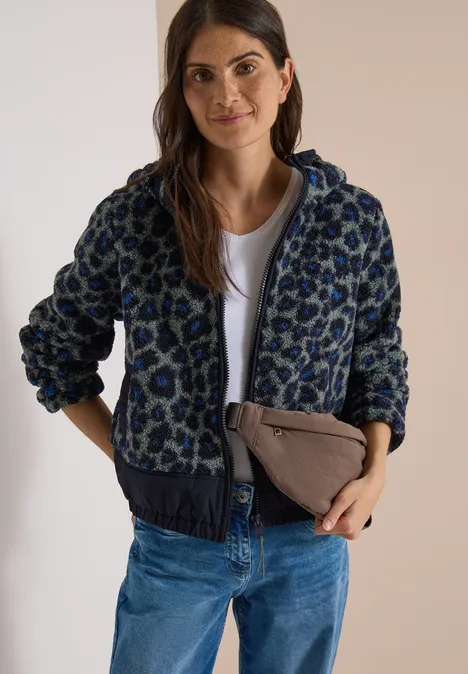 Frau mit gemusterter Jacke, weißem Shirt, Jeans und einer beigefarbenen Clutch lächelt in die Kamera.