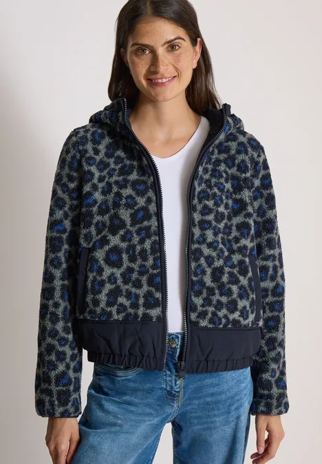 Eine Frau trägt eine dunkelblaue Kapuzenjacke mit Leopardenmuster, weißen T-Shirt und blauen Jeans.