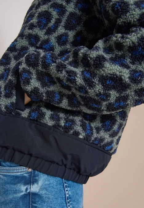 Detailaufnahme einer flauschigen, blau-grünen Leopardenmuster-Jacke mit elastischem Saum über Jeans.