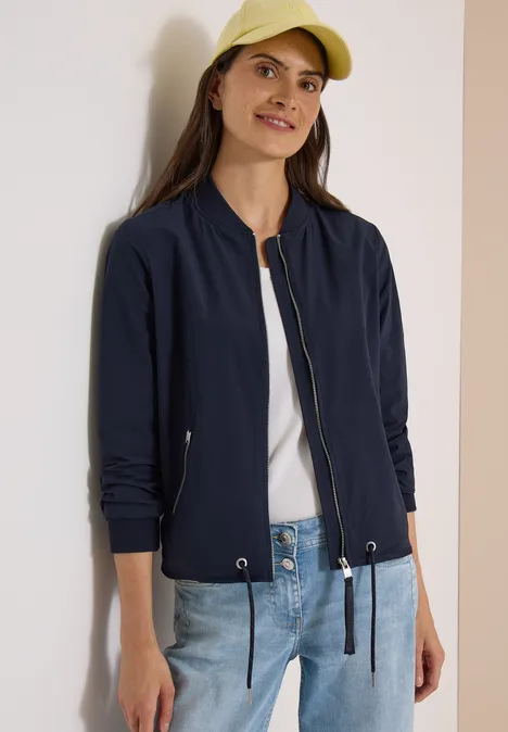 Frau in blauem Bomberjacke, weißem Shirt, Jeans und gelber Cap lächelt entspannt.