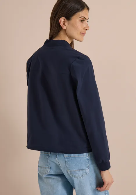 Frau in dunkelblauer, langärmeliger Bluse und hellblauen Jeans, von hinten in natürlicher Pose vor schlichtem Hintergrund.