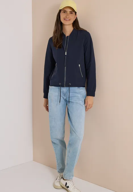 Eine Frau steht an einer Wand, sie trägt eine dunkelblaue Bomberjacke, helle Jeans, weiße Sneaker und ein gelbes Cap.