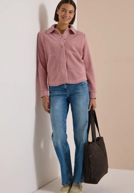 Eine lächelnde Frau lehnt an der Wand, trägt rosa Cordhemd, blaue Jeans und eine große schwarze Tasche.