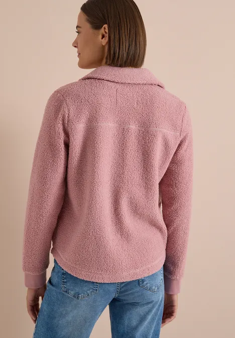 Frau von hinten in rosa Fleecejacke und blauen Jeans vor beigem Hintergrund.