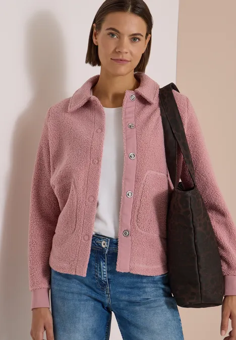 Frau mit rosa Teddy-Jacke, weißem Shirt, blauen Jeans und brauner Tasche vor hellem Hintergrund.