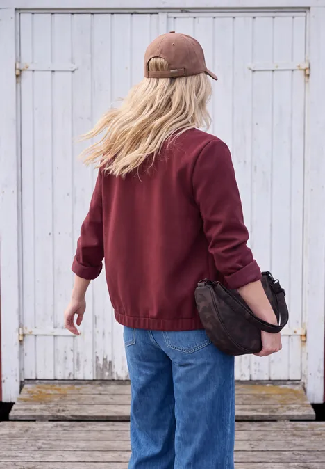 Scuba-piqué collegejasje met ritssluiting red wine Persoon met bordeaux trui, spijkerbroek en pet voor witte houten deuren.