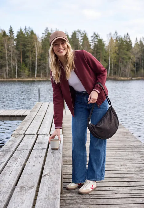 Scuba-piqué collegejasje met ritssluiting red wine Jonge vrouw in jeans, wit shirt, bordeaux jack en pet staat op een houten steiger aan het meer met fles in hand.