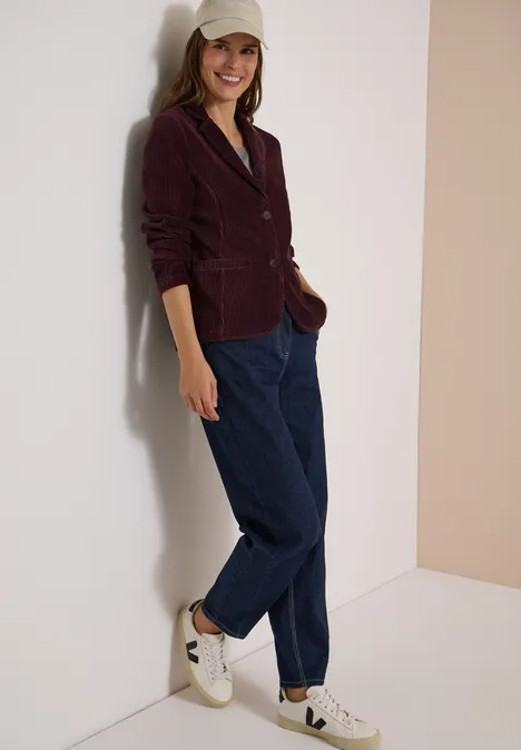 Jerseycord Blazer in Unifarbe berry red Frau lehnt an Wand, trägt burgunderfarbenes Sakko, blaue Hose, weiße Sneaker und eine Baseballkappe und lächelt.