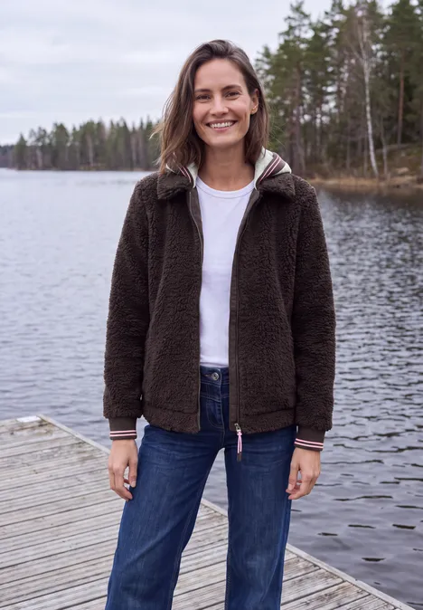 Teddy-Overshirt mit Scuba-Kapuze berry red Frau lächelt auf Steg am ruhigen See, trägt dunkle Jacke, weißes Shirt und Jeans vor Waldkulisse.