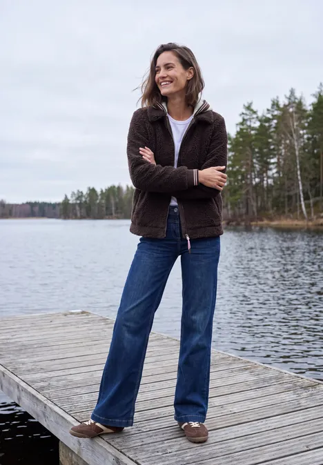 Teddy-Overshirt mit Scuba-Kapuze berry red Eine lächelnde Frau mit brauner Jacke, blauen Jeans und Hausschuhen steht mit verschränkten Armen auf einem Steg am See.