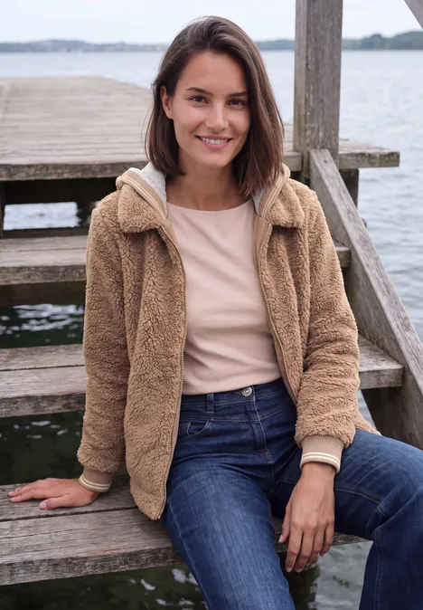 Teddy-overshirt met scuba-capuchon creamy mocha Junge Frau sitzt auf einer Holztreppe am Wasser, lächelt und trägt Jeans und eine kuschelige Jacke.