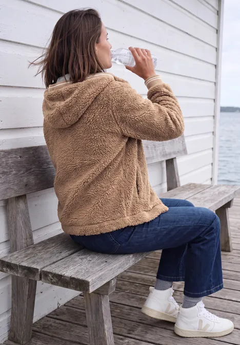 Teddy-overshirt met scuba-capuchon creamy mocha Frau in brauner Teddyjacke und Jeans sitzt auf einer Holzbank vor weißer Hütte am Wasser und trinkt.
