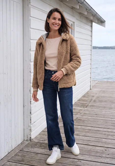 Teddy-overshirt met scuba-capuchon creamy mocha Eine Frau in heller Teddyjacke, Jeans und weißen Sneakern steht lächelnd an einer weißen Holzlaube am See.