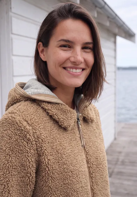 Teddy-overshirt met scuba-capuchon creamy mocha Eine lächelnde junge Frau in braunem Teddy-Mantel steht auf einem Steg am Wasser.