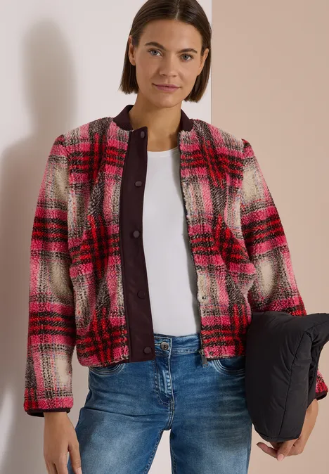 Frau in rotem Karomantel über weißem Top und Jeans, hält schwarze Steppjacke.