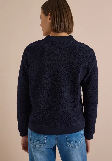 Une personne de dos porte un pull teddy bleu marine devant un fond beige.